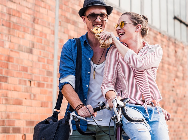 A happy young couple eating pizza and walking with a bicycle