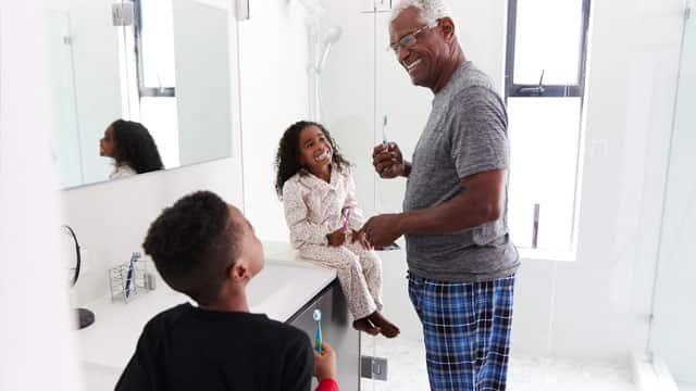 A family of three, that includes an adult and two children, brushing their teeth in front of a mirror.