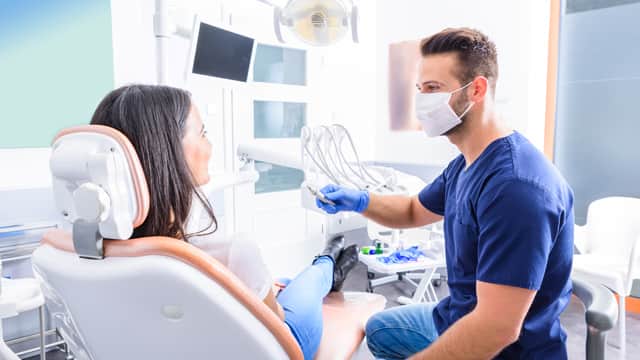 dentist assitant wearing a mask holding a dental tool and a patient sitting