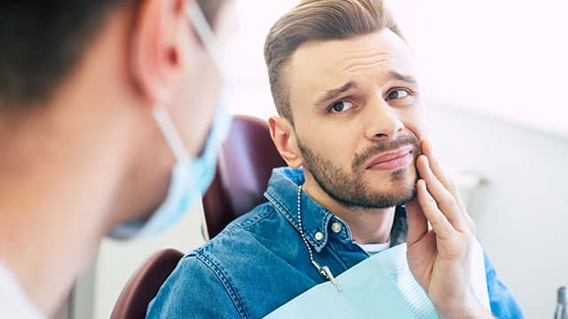 A man with a worried face is holding his hand on his cheek because of irritating pain in front of a dentist