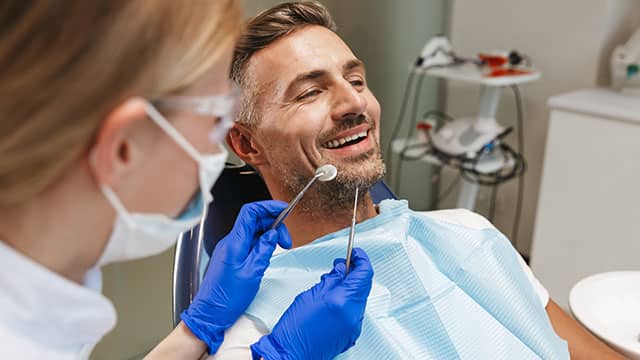 Happy young man sitting in dental chair ready for treatment as a dentist stands next to them holding tools 