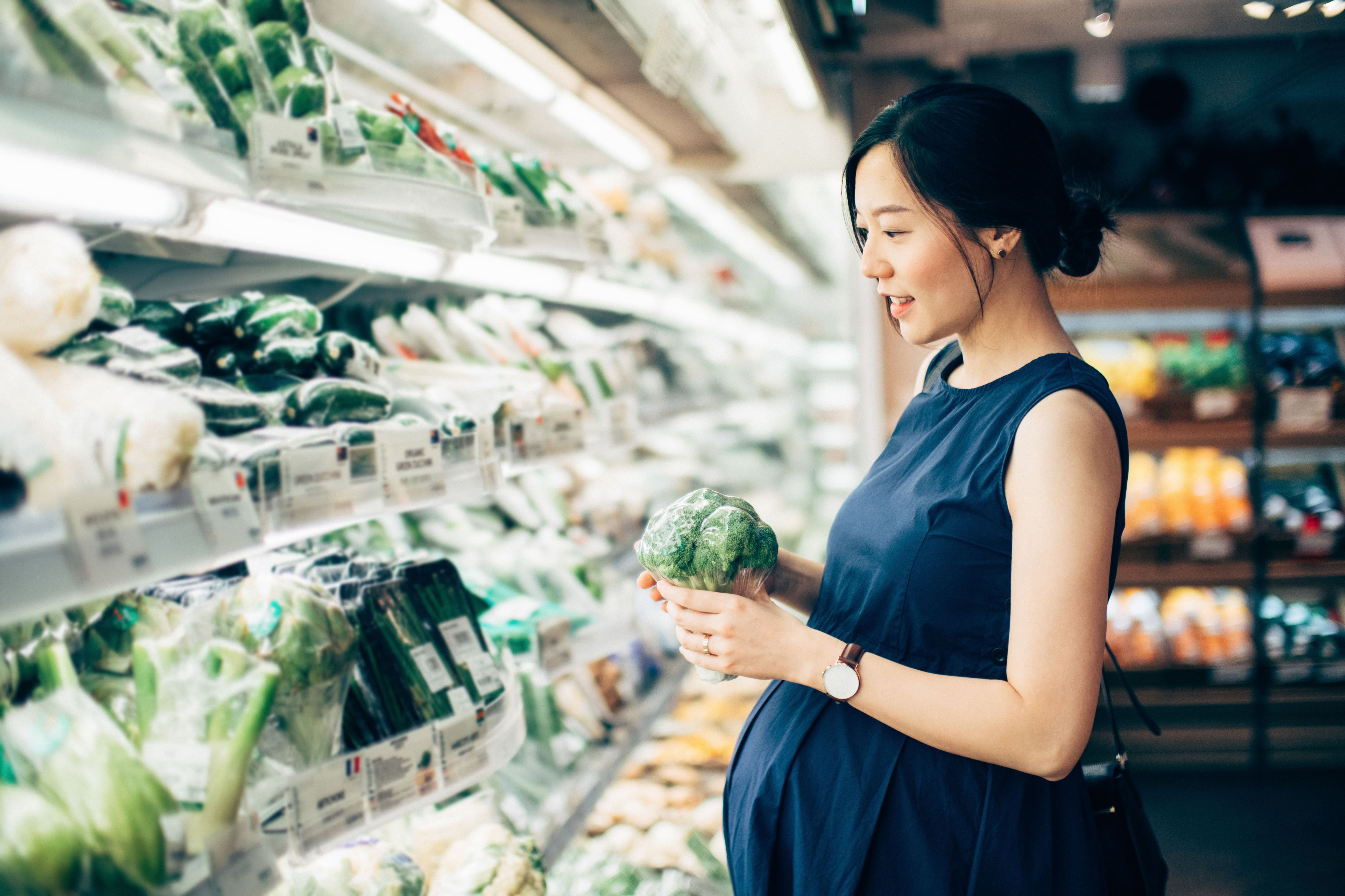 Pregnant woman grocery shopping and looking at the vegetable aisle in a supermarket 