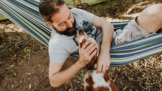 A middle age man in hammock petting a dog outdoors