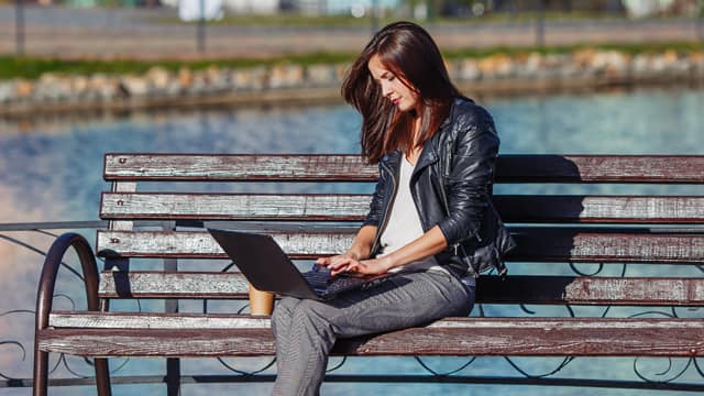 A young woman outdoors on a bench by a pond doing research on a laptop