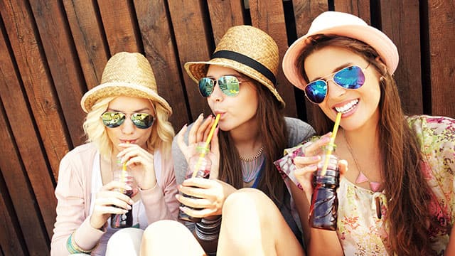 Three young women are drinking soda while wearing sunglasses and hats 