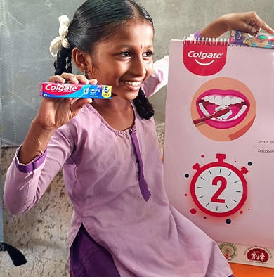 Young girl in India smiling and holding a Colgate Strong Teeth toothpaste box next to an educational oral health poster displaying a two-minute brushing timer.
