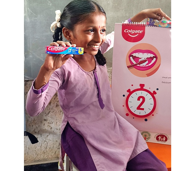 Young girl in India smiling and holding a Colgate Strong Teeth toothpaste box next to an educational oral health poster displaying a two-minute brushing timer.