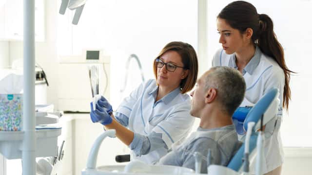 Dentist and her assistant in a dental office talking with a patient and preparing for treatment looking at an x-ray.