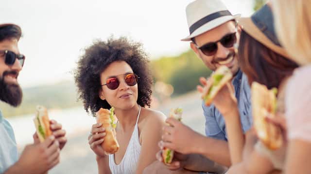 a group of people gathered outdoor smiling while eating a sandwich