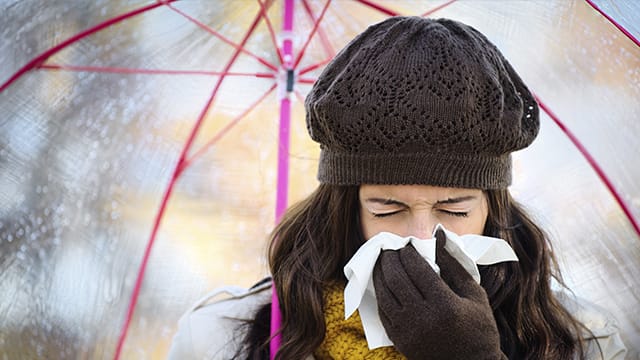  A woman bundled in winter clothing blows their nose under a clear umbrella.