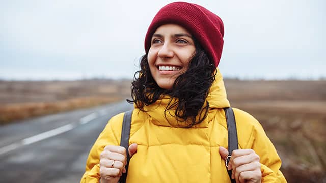 A woman with a backpack wearing yellow jacket and red hat stands on the road
