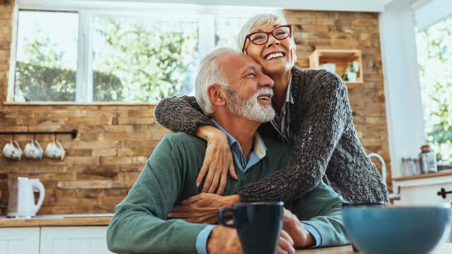 An older woman is hugging her husband in the kitchen