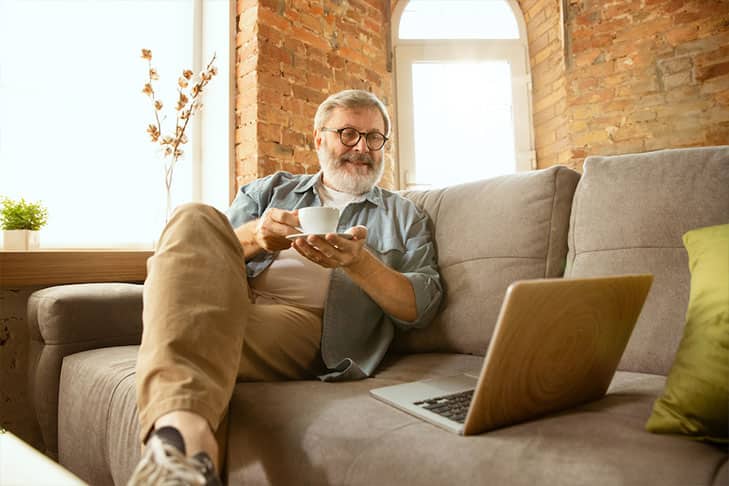 Older man sitting on a couch with a coffee and a laptop 