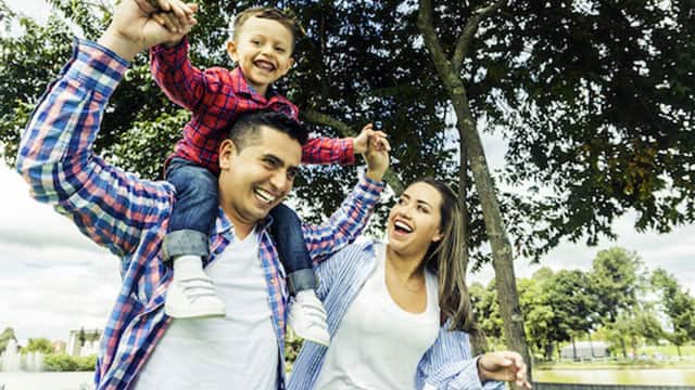 a father smiling holding his kid smiling sitting on his shoulder, and the mother next to them