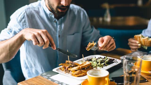 Young man with a beard eating a sandwich with a knife and fork in a restaurant