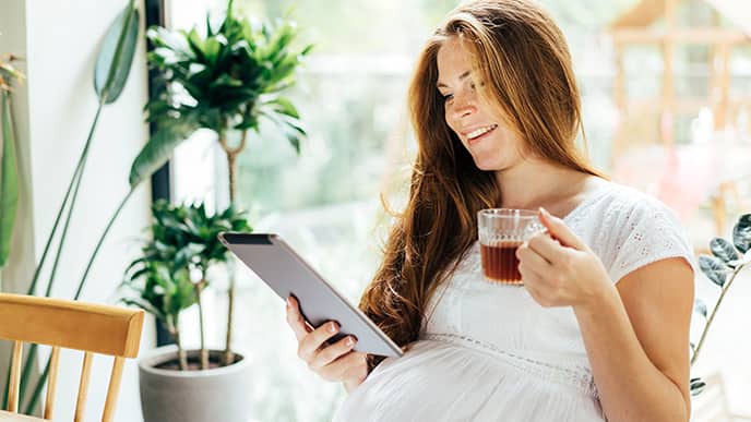 Pregnant woman drinks tea and looks at the screen of a computer tablet.