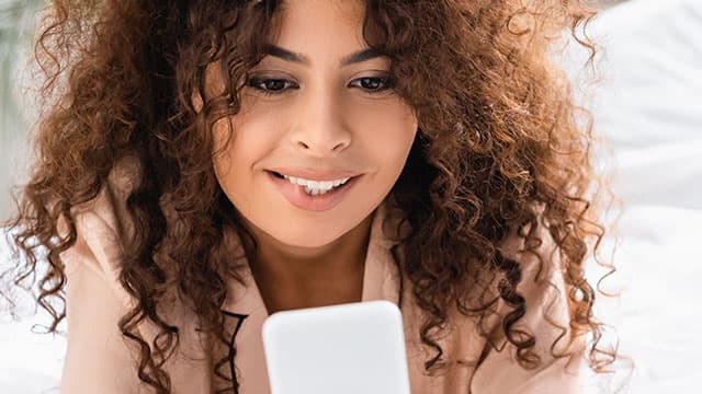 A young woman biting her lip and using smartphone on her bed