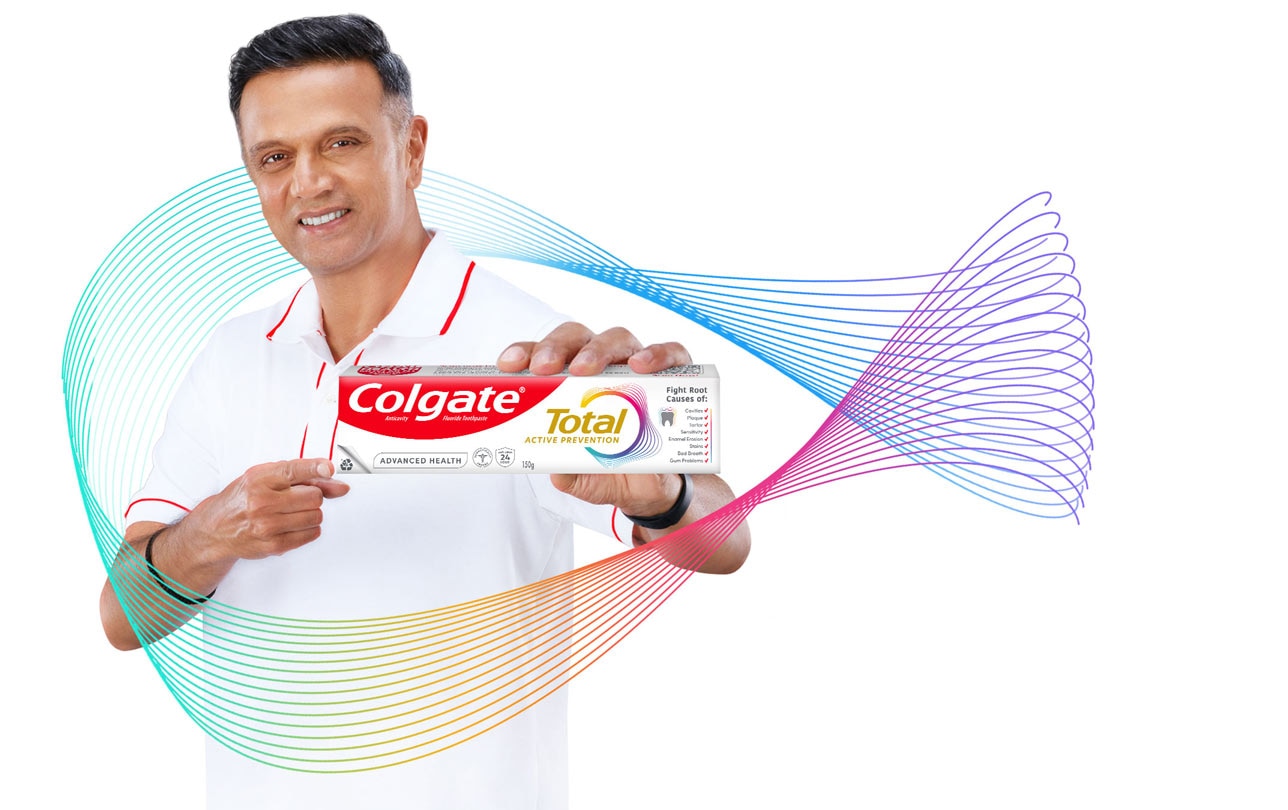 Person smiling while holding and pointing to a box of Colgate Total Active Prevention toothpaste