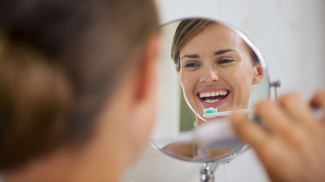 An image of young woman in a mirror brushing her teeth and smiling
