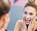 A young woman is looking into a bathroom mirror while touching her jaw and smiling