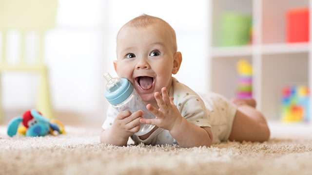 A baby boy is holding a bottle while laying on a bed