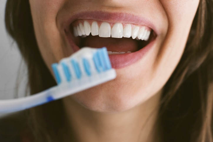 Woman smiles with her mouth open, showing her upper teeth, while holding a toothbrush with toothpaste near her lower lip.