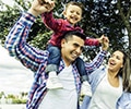 a father smiling holding his kid smiling sitting on his shoulder, and the mother next to them