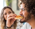 A man with long curly hair and a beard eating a pizza bagel