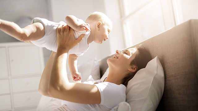 A mother carrying her baby in the air while laying on her back on bed having good time