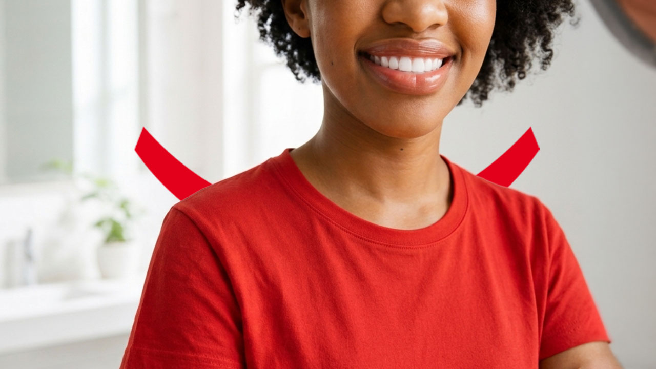 A smiling woman with dark curly hair and a red t-shirt standing in an all-white bathroom. 
