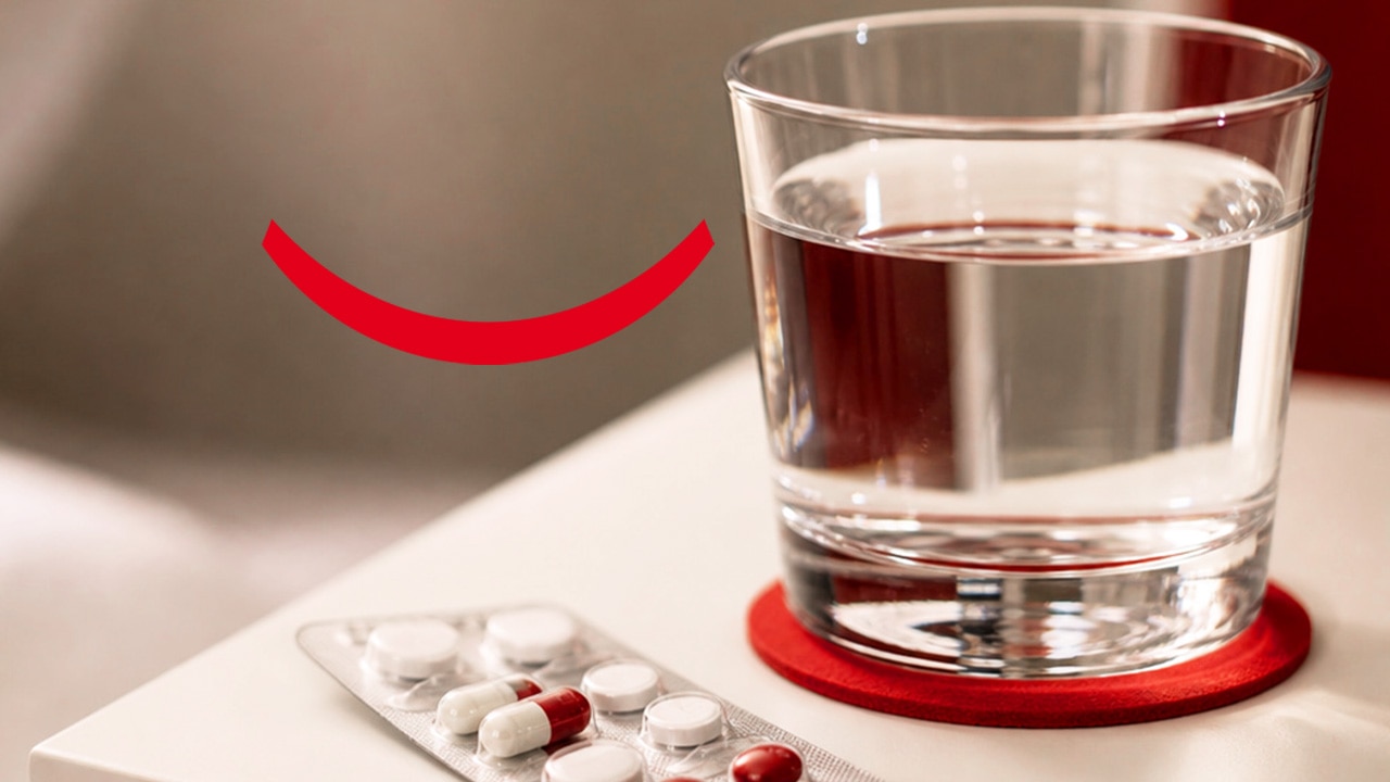 A glass of water on a nightstand with a red coaster next to a blister pack of heartburn tablets and other medications.  