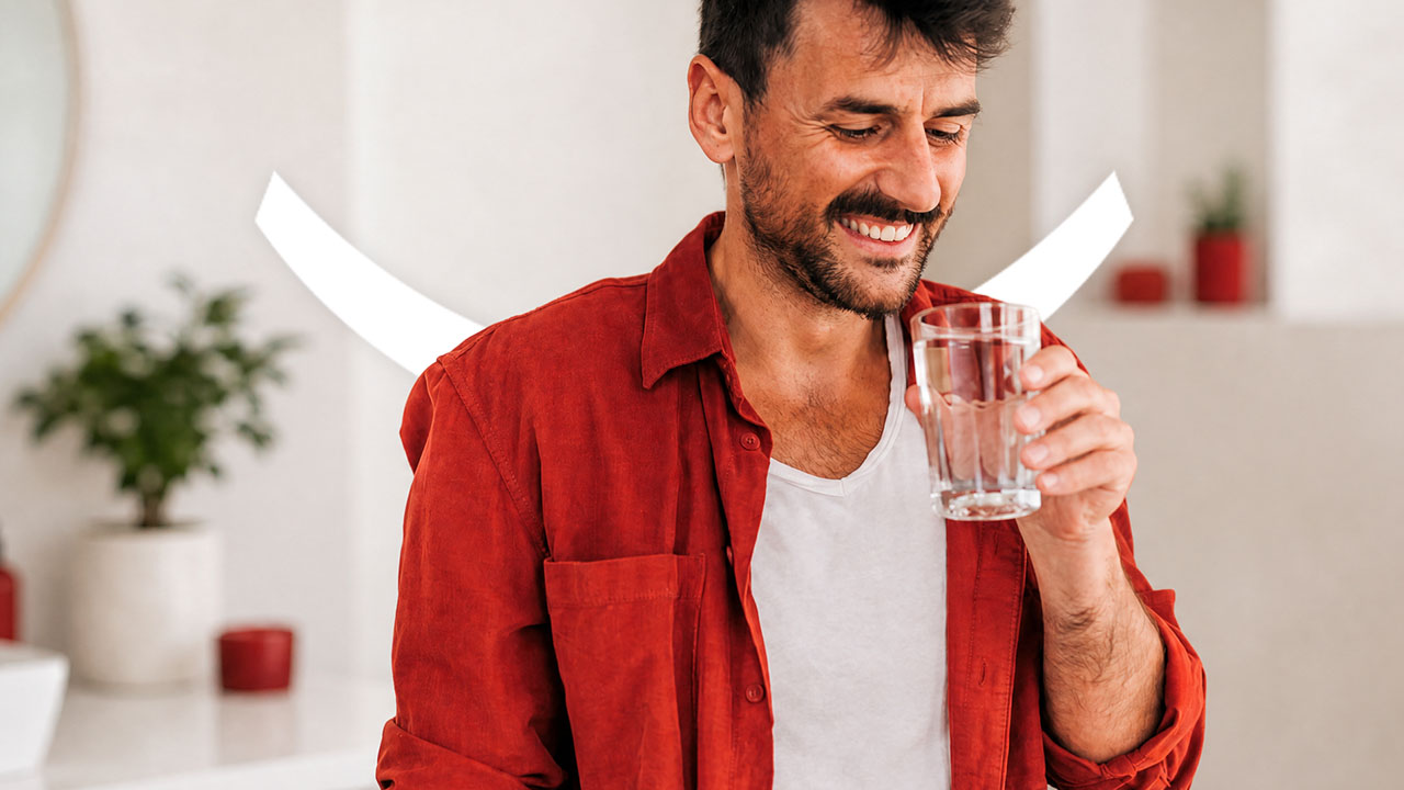 A smiling, dark-haired man in a white vest and red shirt, holding a glass of water up to his mouth to relieve GLP-1 dry mouth. 