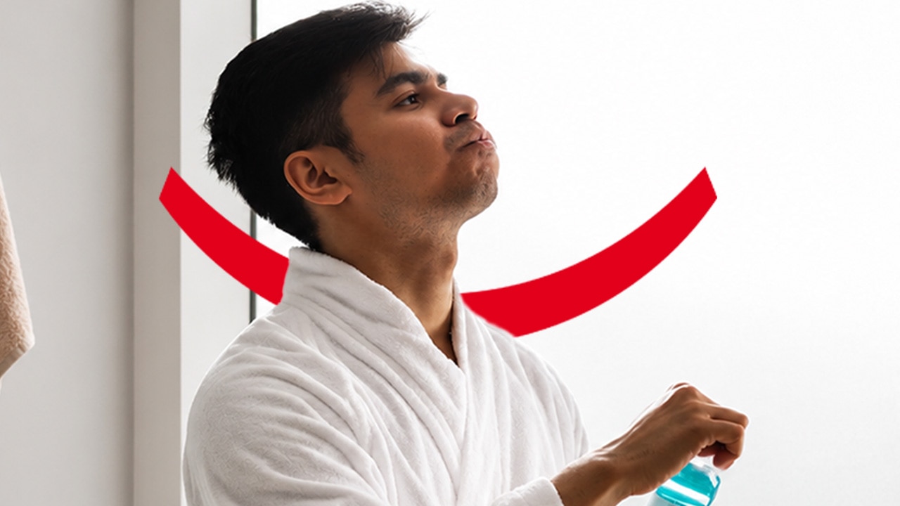 A dark-haired man in a white bathrobe holding a bottle of Colgate mouthwash and swishing some around his mouth with his head tilted back. 