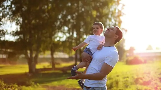A smiling man outside, holding a child while looking up