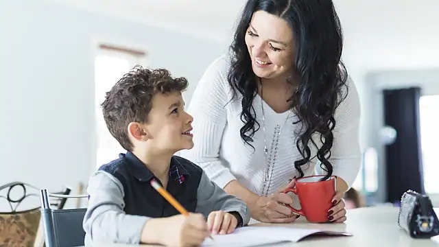 A woman smiling at a young boy doing his homework