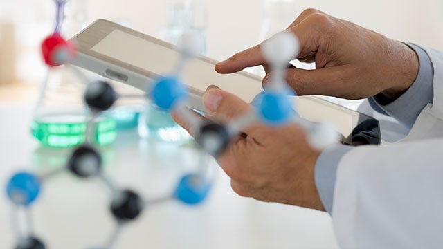 Close-up of hands holding digital tablet near a molecular model and lab flasks in the background