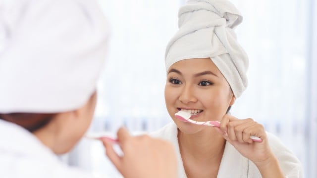Close-up of young women brushing her teeth in front of the mirror in her robe and with a head towel on 