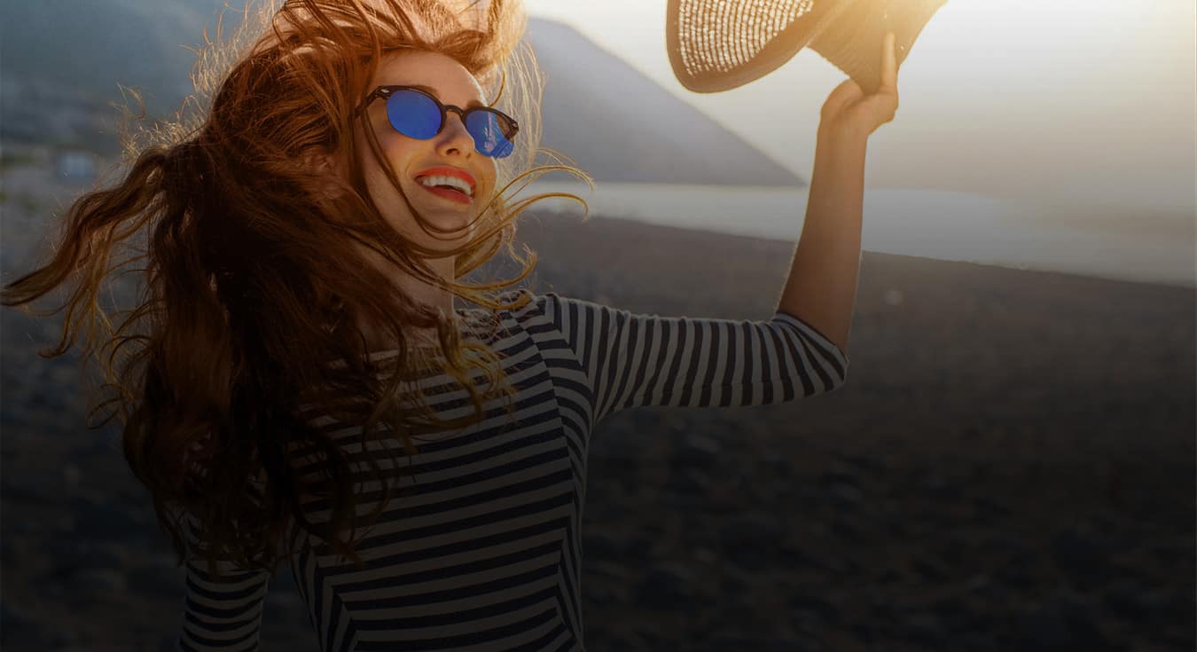 woman wearing sunglasses smiling at the beach holding a hat