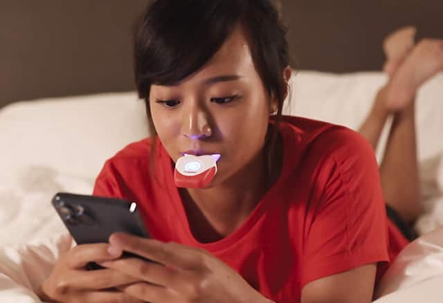 Lady using Colgate's teeth whitening kit while she uses her phone on the bed