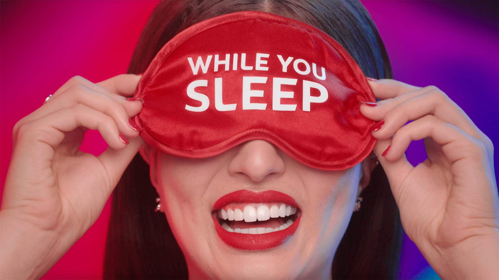 Ann smiling loudly while holding on a eye mask with the word while you sleep on it