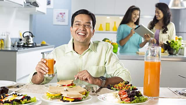 a man smiling while holding a glass of juice and a mother and daughter in the back