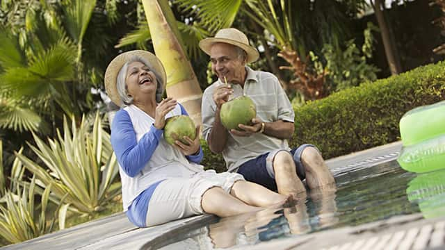 an elderly couple smiling sitting down at the pool while enjoying a coconut drink