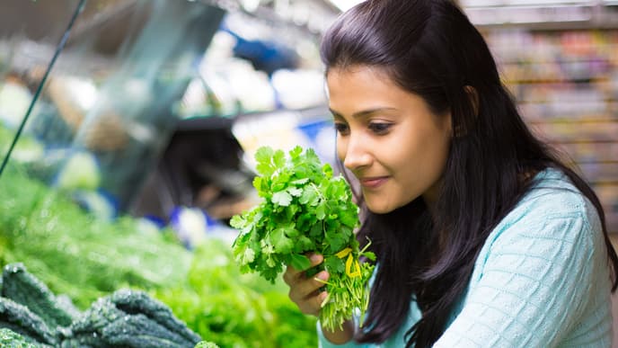 Closeup portrait, beautiful, pretty young woman in sweater picking up, smelling, choosing green leafy vegetables in grocery store