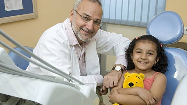a girl sitting on the dental chair smiling hugging a teddy bear next to the smiling dentist