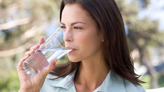 a young women in light blue shirt is drinking water from a glass outdoors