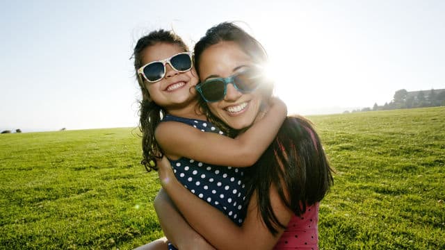 Açık havada güneş gözlüğü takan bir anne ve kızı birbirlerine sarılırken parlak bir şekilde gülümsüyorlar.