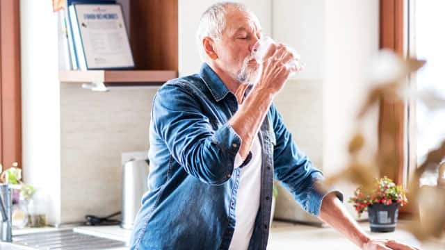 An older gentleman drinking water in a kitchen.