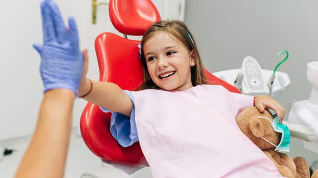 Little girl at dentist holding a teddy bear and high fiving the dentist.