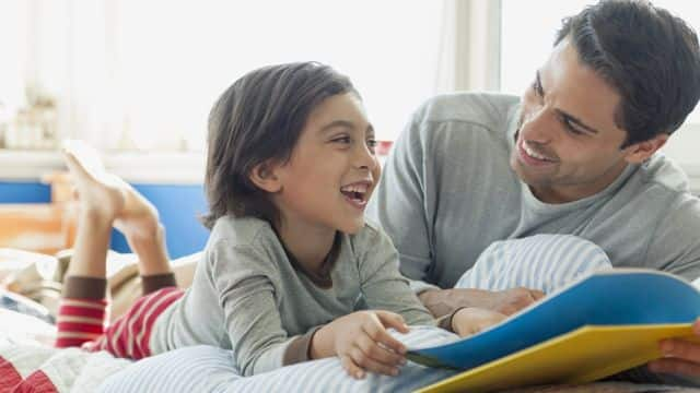 a father smiling laying down next to his son reading a book smiling