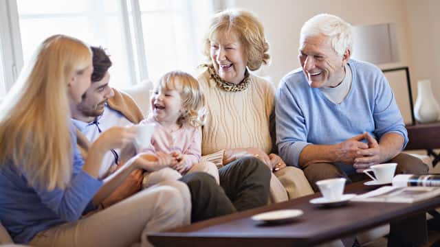 a grandfather, grandmother, mother, father and a baby girl smiling brightly while sitting on the couch